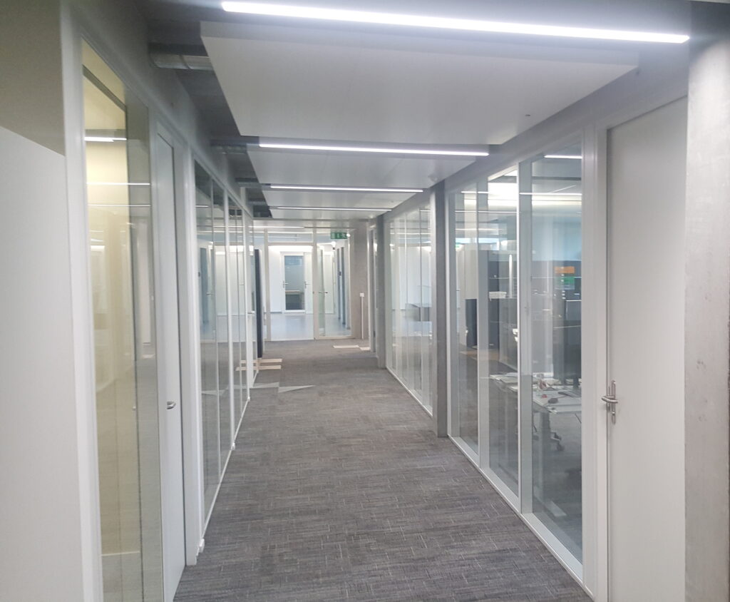 Moderner Büroflur mit Glastrennwänden, grauen Teppichfliesen und LED-Deckenbeleuchtung