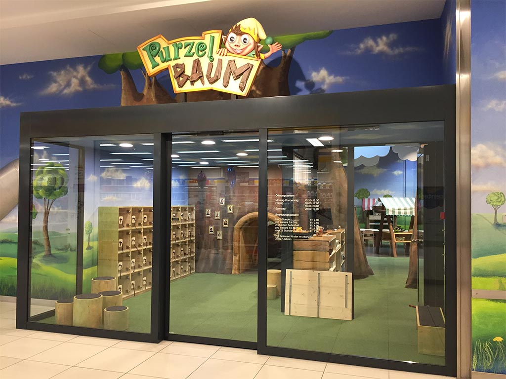 Eingang zum Kinderspielbereich Purzelbaum mit Glasfront, Holzmöbeln und bunter Waldkulisse in einem Einkaufszentrum