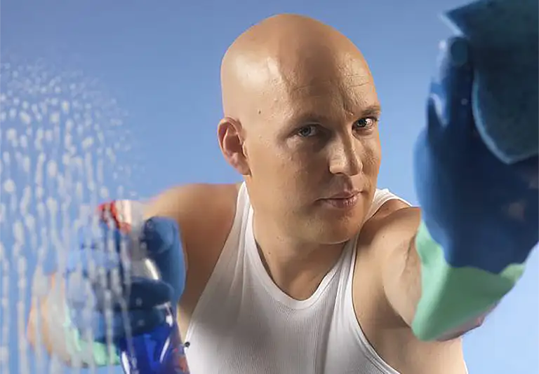 Glatzköpfiger Mann im weißen Unterhemd putzt mit Sprühflasche und Tuch eine Glasfläche