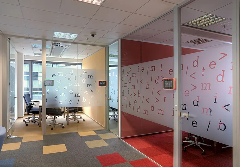 Moderne Büroräume mit Glastrennwänden, dekorativen Buchstaben und farbigen Teppichfliesen in Grau, Gelb und Rot