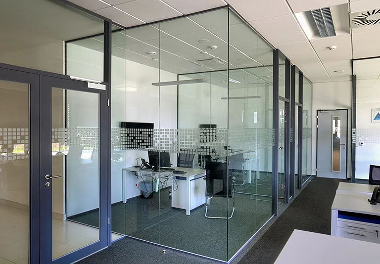 Modernes Büro mit Glastrennwänden, dunklen Aluminiumrahmen, weißen Schreibtischen und Computerarbeitsplätzen