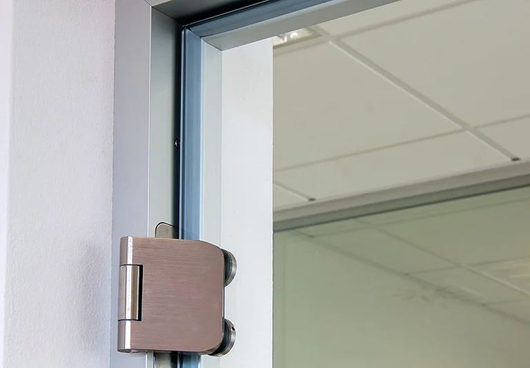 Glastür mit gebürstetem Edelstahl-Scharnier an weißer Wand, Blick auf Deckenplatten im Hintergrund