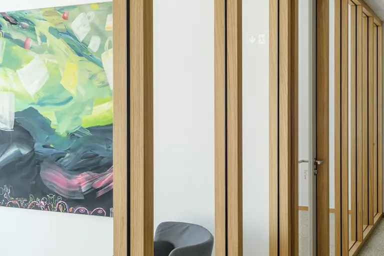 Moderner Büroflur mit Holz-Glas-Trennwand, abstraktem Gemälde in Grün-Gelb-Tönen und grauem Sessel