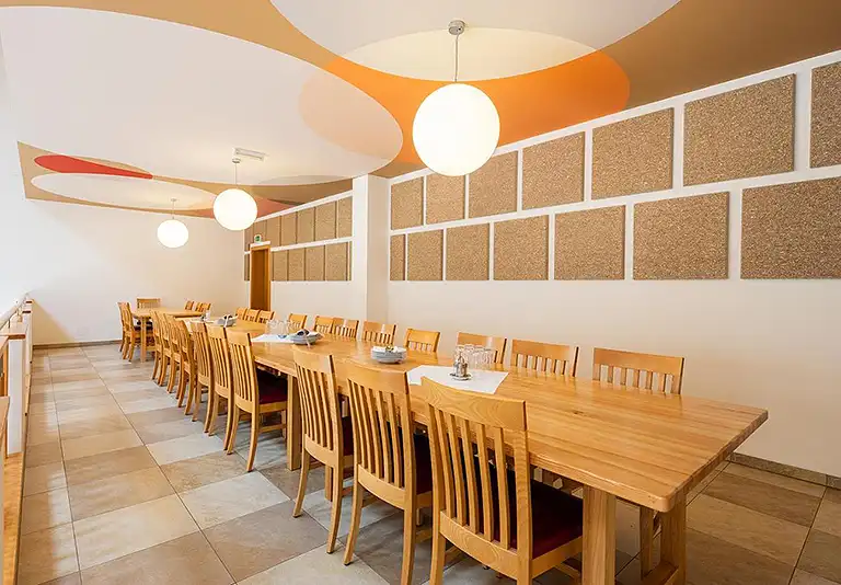 Heller Speisesaal mit langen Holztischen, Stühlen und runden Deckenlampen, Wände mit Korkpaneelen und orangefarbenen Akzenten
