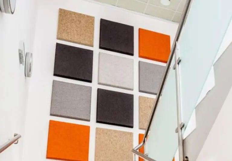 Quadratische Akustikpaneele in Orange, Grau, Schwarz und Kork an weißer Treppenhauswand mit Glasgeländer