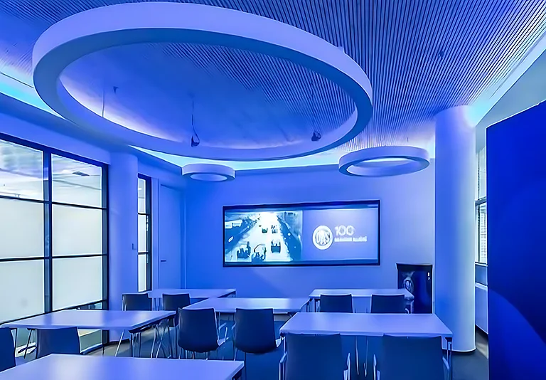 Moderner Konferenzraum mit blauer LED-Beleuchtung, weißen Tischen, dunklen Stühlen und großer Leinwand