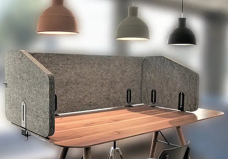 Schreibtisch aus Holz mit grauem Filz-Sichtschutz an drei Seiten, darüber drei Hängelampen in verschiedenen Farben