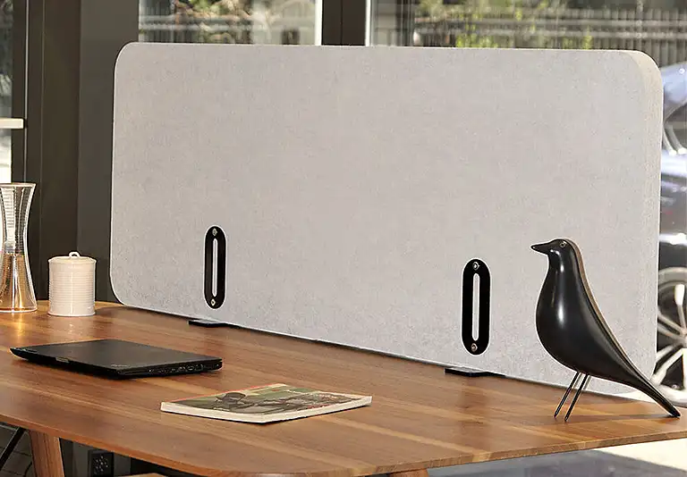 Grauer Akustik-Tischaufsatz mit schwarzen Klemmen auf Holzschreibtisch, daneben Laptop, Eames-Vogelfigur und Vase