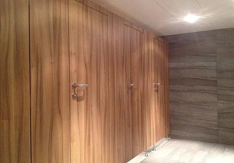 Drei WC-Kabinen mit eleganten Holztüren in warmem Nussbaumton, silberne Türgriffe, graue Steinwand rechts