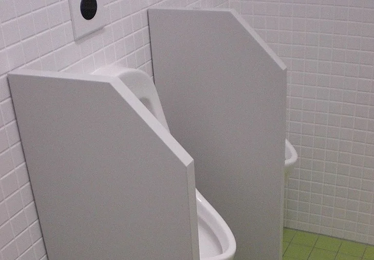 Zwei weiße Urinale mit Trennwänden in einem gefliesten Sanitärraum mit weißen Wandfliesen und grünem Boden